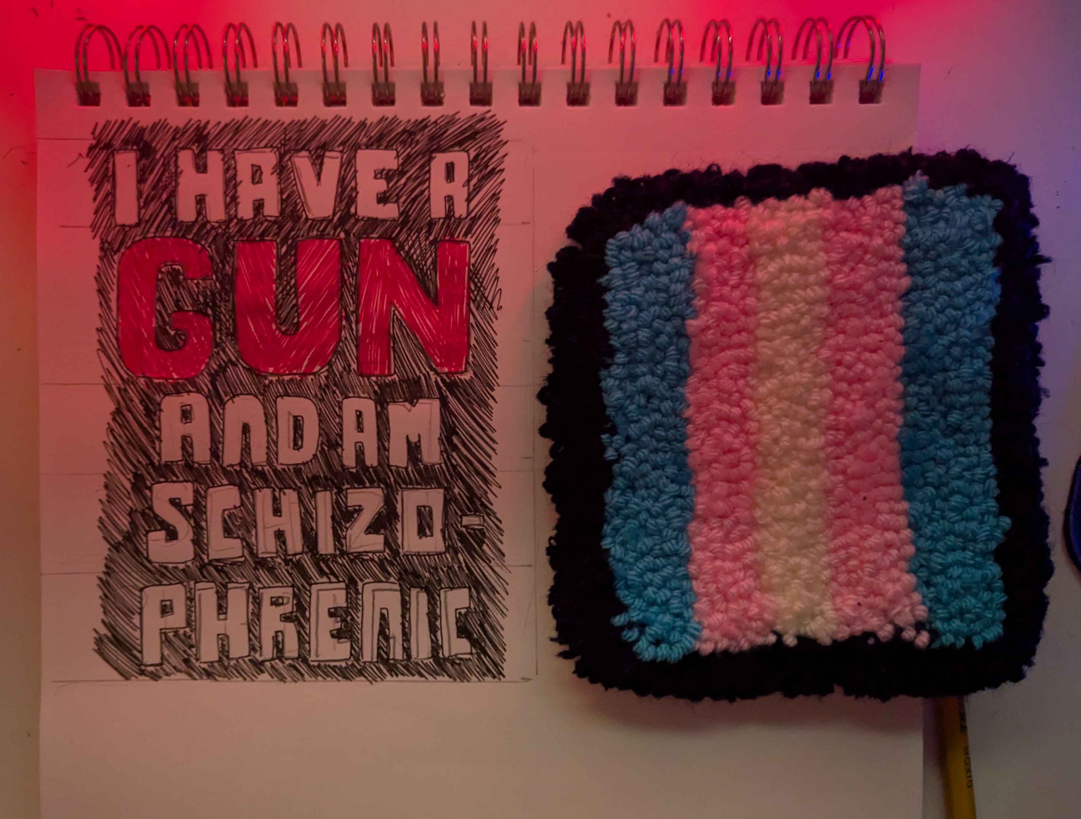 sketch concept for a rug. The text reads 'I have a gun and am schizophrenic'. It is pictured next to a completed trans flag rug, for scale